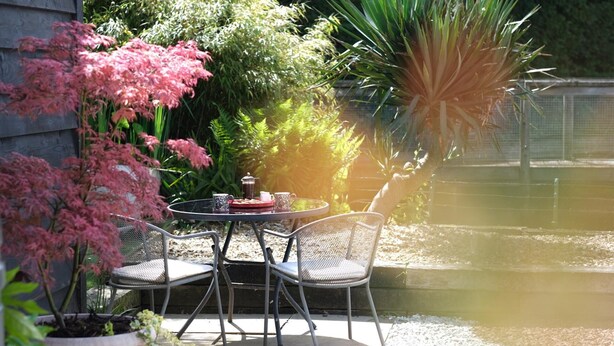 Une terrasse extérieure avec une table ronde et deux chaises, entourée de plantes en pot et d’un petit bassin.