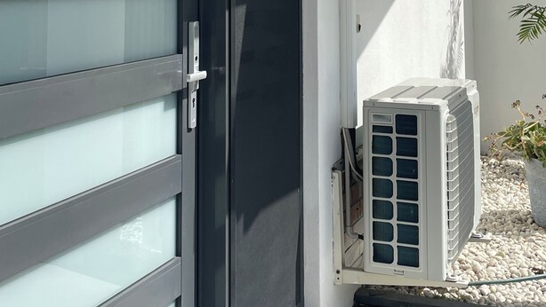 A modern building with a sliding glass door, a white exterior wall, and an air conditioning unit on the ground.