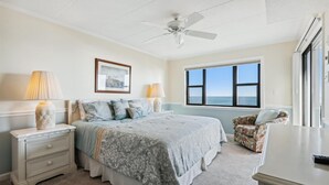 A bedroom with a bed, bedside lamps, a ceiling fan, a framed picture, a window with a view, and a chair. - Enjoy beautiful ocean views at this 3 bedroom, 2 bath oceanfront condo located downtown! (Ocean City)