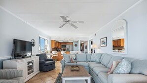 A spacious living room with a large grey sofa, a wooden coffee table, and a television on a cabinet. There is a ceiling fan, a kitchen visible in the background, and a large mirror on the wall. - Enjoy beautiful ocean views at this 3 bedroom, 2 bath oceanfront condo located downtown! (Ocean City)