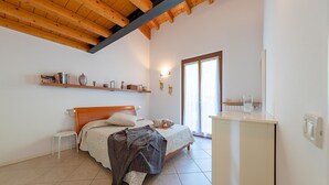 Una camera da letto con soffitto in legno, un letto con copripiumino bianco, un comodino, una piccola sedia bianca, un altro comodino in legno e una finestra con tende.