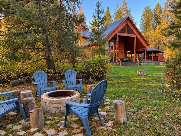 A cabin with a stone fire pit and blue Adirondack chairs in a forest setting.