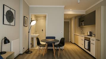 A compact hotel room with a bed, bedside table, lamp, and a dining area with chairs and a table.