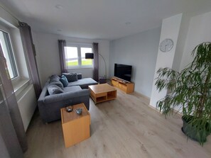 Ein modernes Wohnzimmer mit grauem Sofa, einem Holztisch, einem Fernseher auf einem Holzgestell und einer großen Topfpflanze.