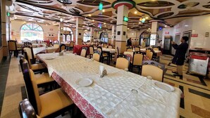 A spacious dining area with long tables and chairs, a high ceiling with unique architectural details, and a large window providing natural light. - Hotel Sea World Cox's Bazar (Cox's Bazar)