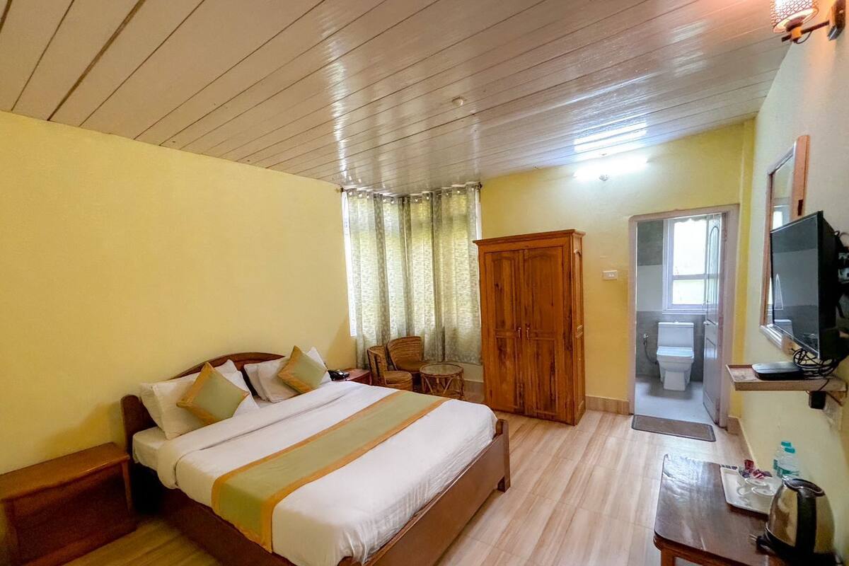 Una habitación de hotel con una cama de madera, un armario de madera, una mesita con un hervidor, un televisor y un baño visible a través de una puerta abierta.