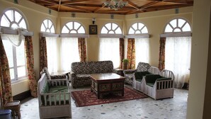 Une pièce avec un canapé à motifs, une petite table et des chaises en osier. De grandes fenêtres sont présentes, ornées de rideaux.