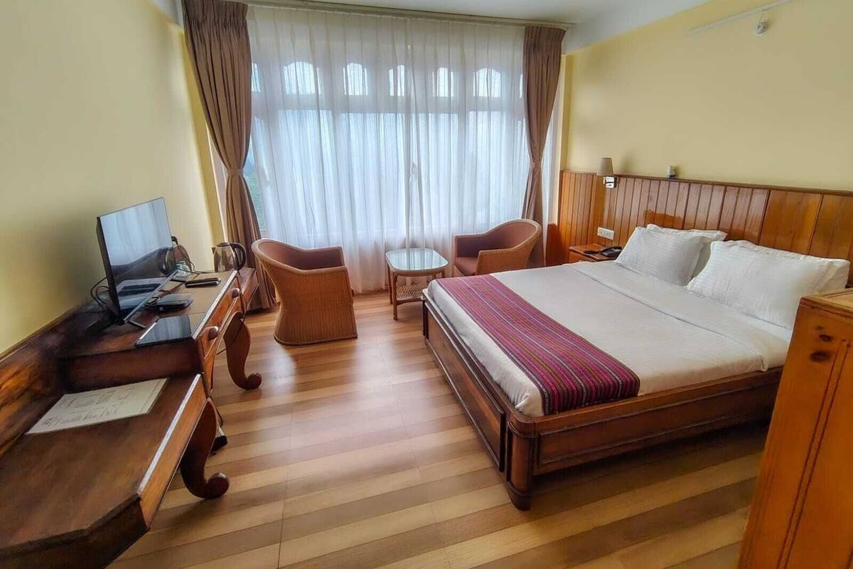 Habitación de hotel con una cama de madera, un escritorio con computadora, dos sillas y una ventana con cortinas.