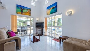 Amplia sala de estar con sofá, mesa de centro y televisor, ventanas grandes y obras de arte en las paredes.
