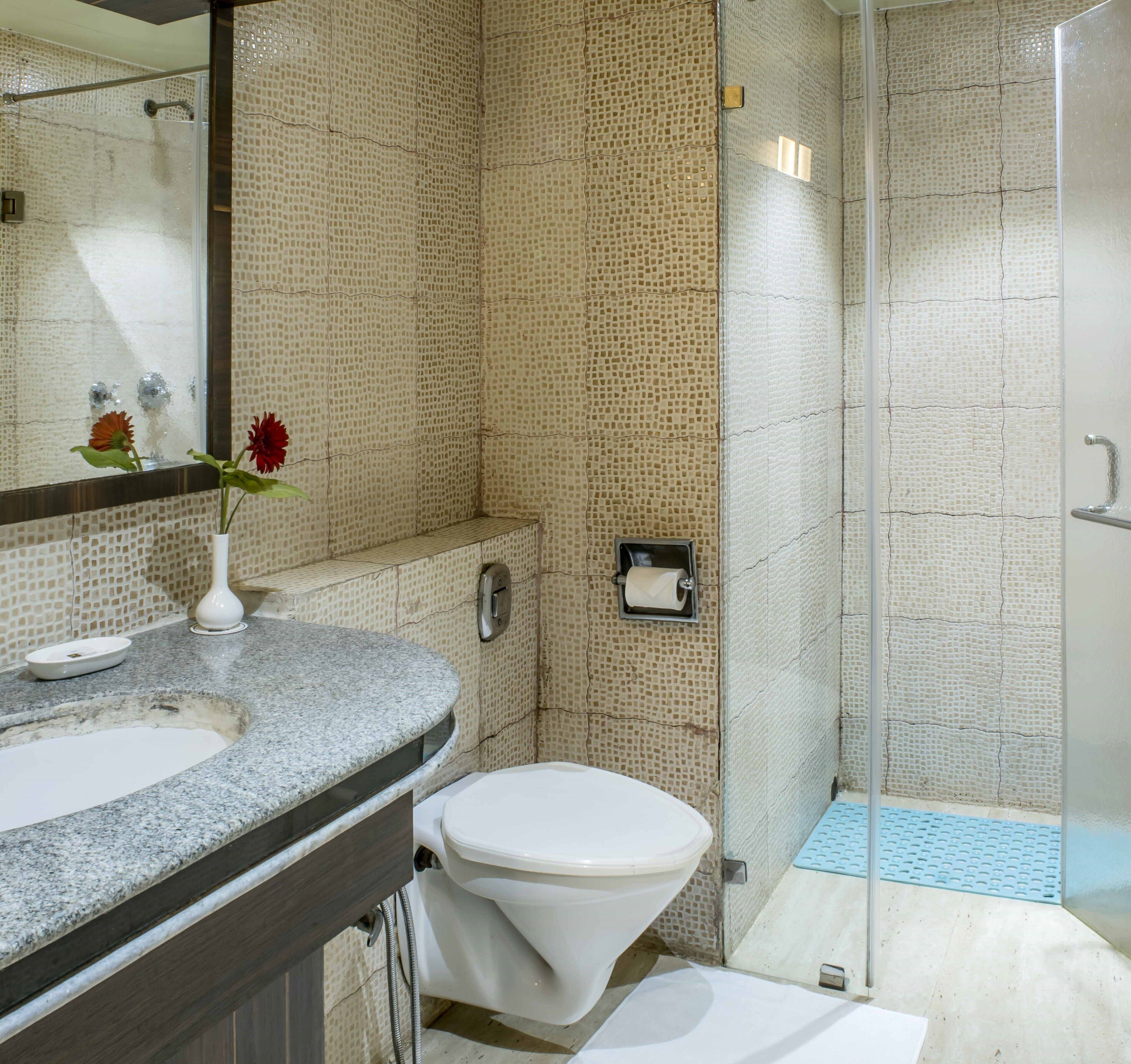A bathroom with a granite countertop, a white toilet, a glass shower enclosure, and a mirror with a vase of flowers.