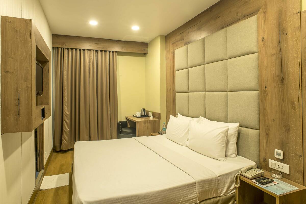 Quarto de hotel com uma cama grande, cabeceira de madeira, escrivaninha com cadeira, mesa pequena com abajur e janela com cortinas.