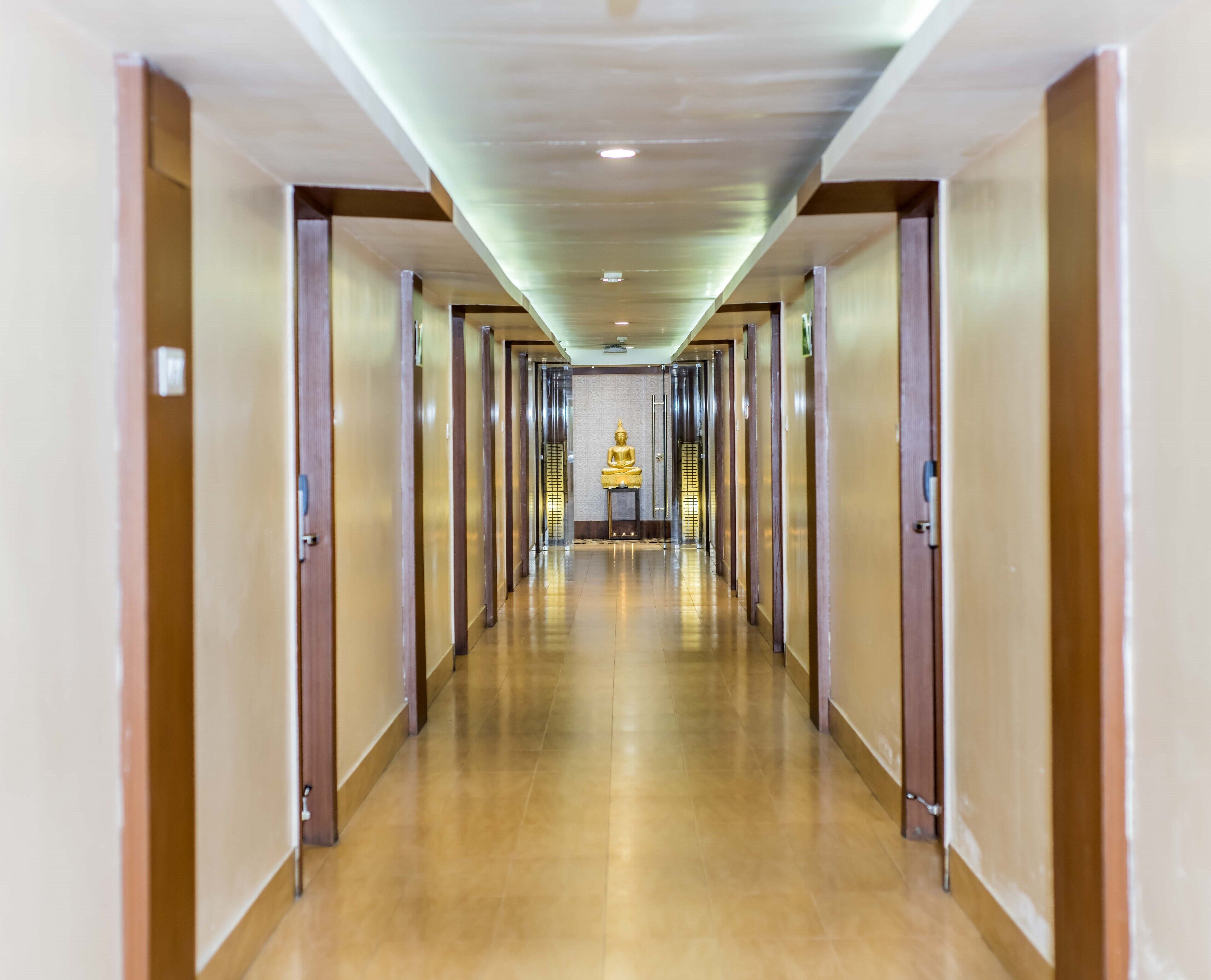 Un largo y estrecho pasillo de hotel con puertas de madera a ambos lados y una estatua dorada de Buda al final.