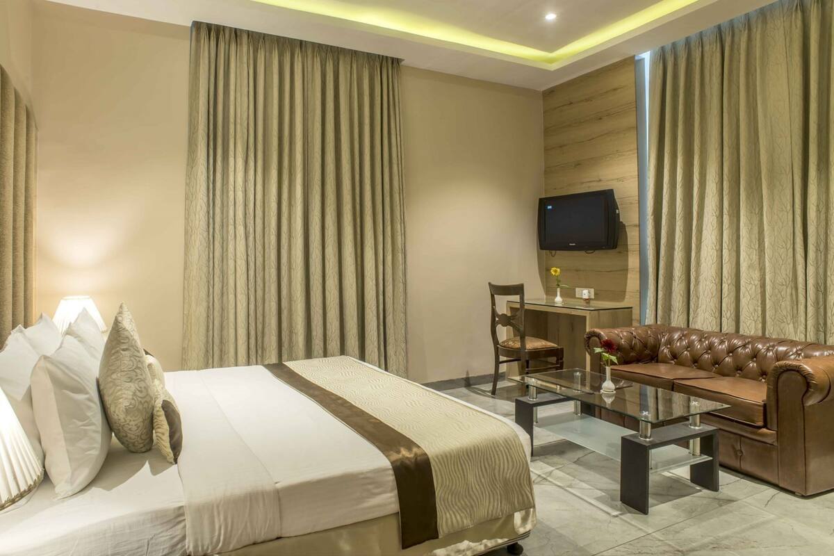 Quarto de hotel com cama, sofá de couro, mesa de centro de vidro, escrivaninha e televisão na parede.