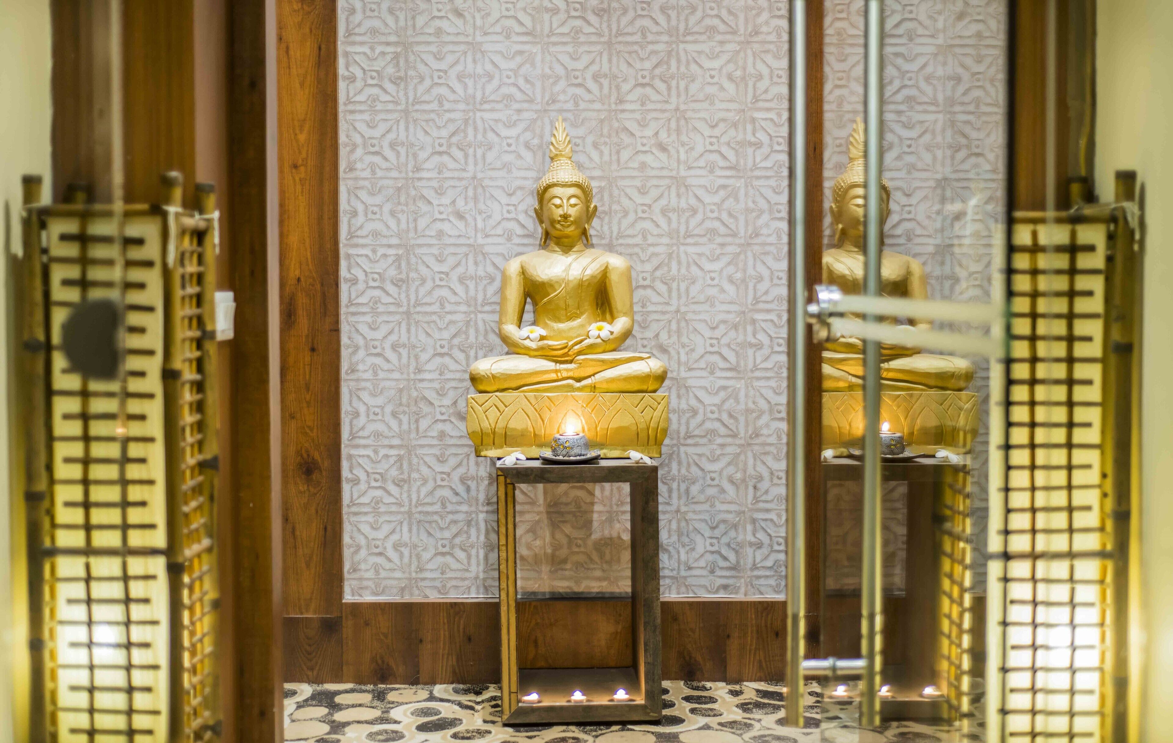Uma estátua dourada de Buda sobre um pedestal, com velas, diante de uma parede com padrão.