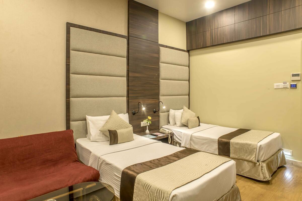Quarto de hotel com duas camas, um sofá, uma mesinha de cabeceira e um candeeiro fixo na parede.