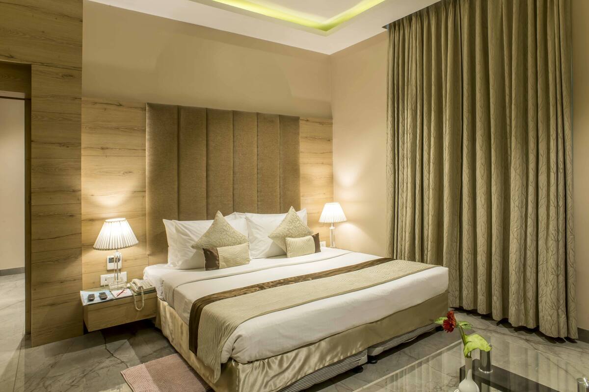 Quarto de hotel com uma cama grande, duas luminárias de cabeceira, uma mesa de cabeceira com telefone e um vaso com uma flor no chão.