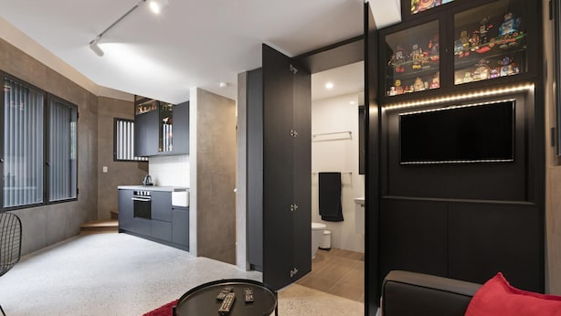 Un salon moderne avec un canapé noir, un tapis rouge qui apporte une touche de couleur, et un téléviseur fixé au mur.