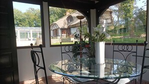 Mesa de vidrio con sillas de metal en un espacio exterior cubierto, con vista a una casa y árboles.