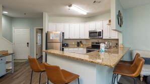 Una cocina moderna con armarios blancos, encimeras de granito y electrodomésticos de acero inoxidable. Hay dos taburetes de bar y una pequeña isla con un fregadero.