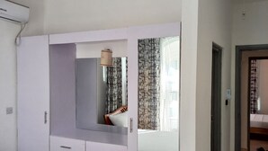 A bedroom with a bed, a wardrobe with a mirror, a ceiling fan, and a window with patterned curtains. - Kijani Homes Apartments Ocean Beach Drive (Malindi)