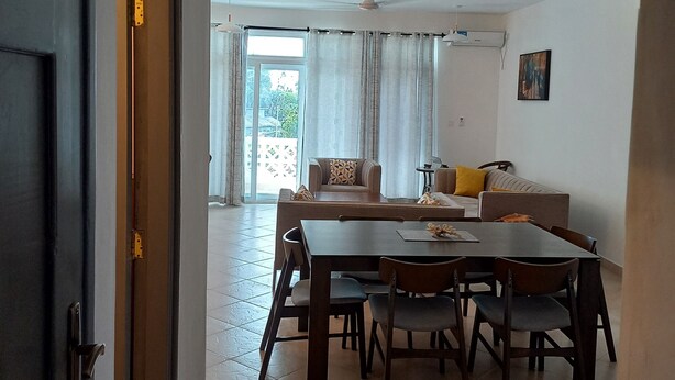 A dining area with a wooden table and chairs, a ceiling fan, and a view of the outdoors through large windows. - Kijani Homes Apartments Ocean Beach Drive (Malindi)
