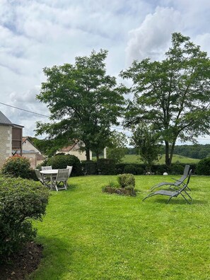A grassy outdoor area with a table and chairs, a small garden, and a building in the background.
