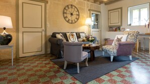Een gezellige woonkamer met een zwart-wit betegelde vloer, een bank, twee fauteuils, een salontafel, een wandklok en een raam met natuurlijk licht.