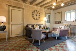 Un salon chaleureux avec un sol à carreaux, un canapé, deux fauteuils, une table basse, une horloge murale et une fenêtre laissant entrer la lumière naturelle.