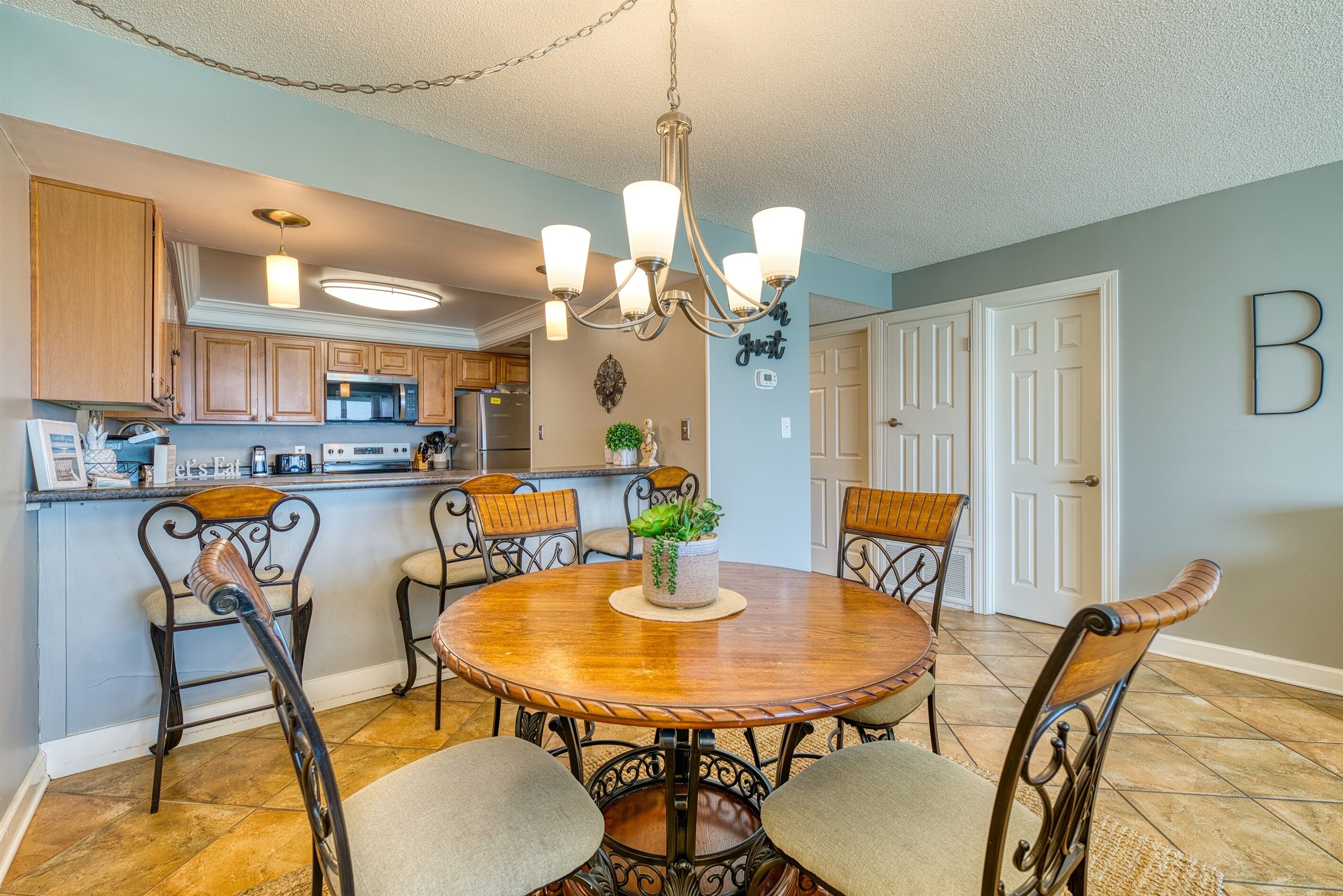 A dining area with a round wooden table, metal chairs, a chandelier, and a kitchen in the background.
