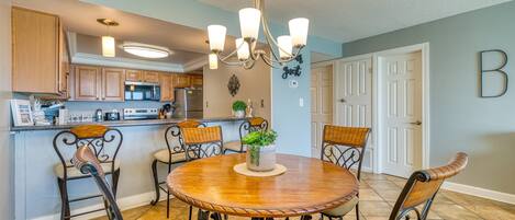 A dining area with a round wooden table, metal chairs, a chandelier, and a kitchen in the background.