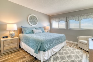 A bedroom with a bed, two bedside tables, a wall clock, a decorative wall piece, lace window valances, and a patterned rug.