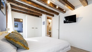Une chambre avec un grand lit, des poutres en bois au plafond, un téléviseur à écran plat et une salle de bain visible à travers une porte ouverte.
