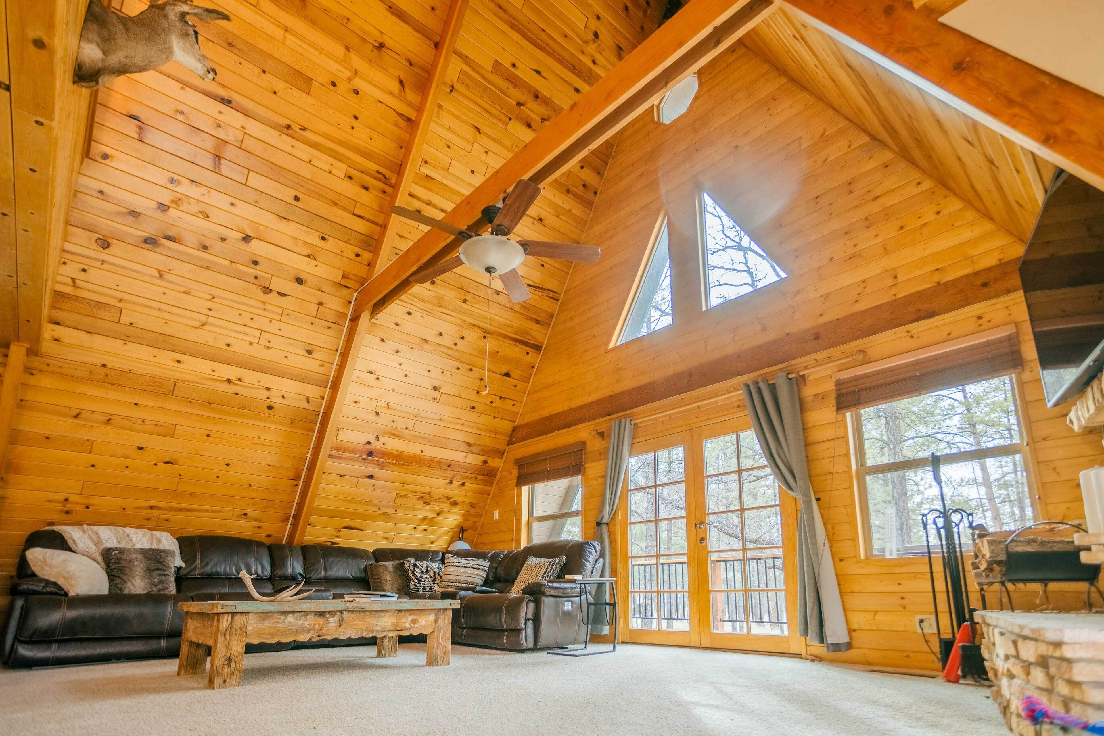A log cabin interior with a wooden ceiling, a ceiling fan, a leather sofa, a coffee table, and large windows.