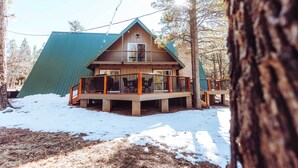 A cabin with a green roof, wooden deck, and a balcony with chairs. - Furnished Deck + Fire Pit: Happy Jack Cabin (Happy Jack)