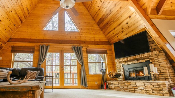 A log cabin interior with a stone fireplace, a ceiling fan, and a large window. - Furnished Deck + Fire Pit: Happy Jack Cabin (Happy Jack)