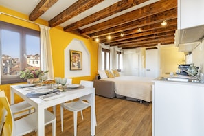 A room with a wooden ceiling, a bed, a dining table with chairs, and a view of buildings outside.