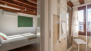 Ein Schlafzimmer mit Bett, Wand mit grünem Muster und Holzboden. Es zeigt außerdem ein Badezimmer mit Glasdusche, Handtuchhalter und Blick ins Freie.