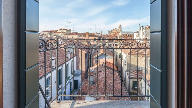 Blick von einem Balkon mit schmiedeeisernem Geländer auf eine Stadtansicht mit Häusern mit Terrakotta-Dächern und einem klaren Himmel.