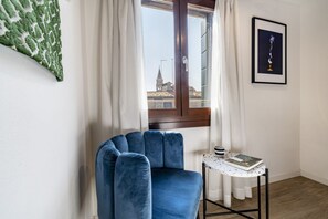 Habitación de esquina con una silla azul, una mesita con un libro, un cuadro en la pared y una ventana con cortinas.