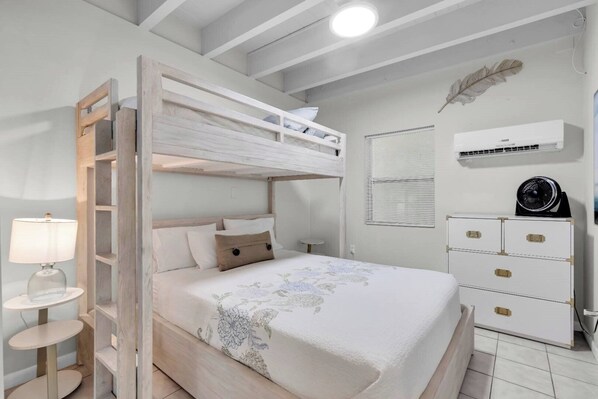 Un lit superposé avec une tête de lit blanche, une table de chevet avec une lampe, une commode avec des tiroirs et un appareil de climatisation fixé au mur.
