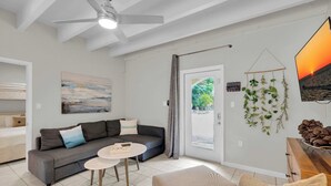 Un salon avec un canapé gris, une table basse ronde, un fauteuil beige, un ventilateur de plafond, un tableau, un téléviseur et une porte donnant sur un paysage verdoyant.