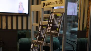 A reception area with a sign reading "Grand Ulger Hotel," a magazine rack with 'ERCIYES' magazines, a green sofa, and a television displaying a blurred image.