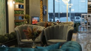 A seating area with upholstered chairs and a vase of dried flowers, a fireplace, and a view of parked cars through large windows. - Grand Zara Otel (Kayseri)