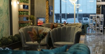 Un área de estar con sillones tapizados, un jarrón de flores secas, una chimenea y vistas a coches estacionados a través de amplias ventanas.