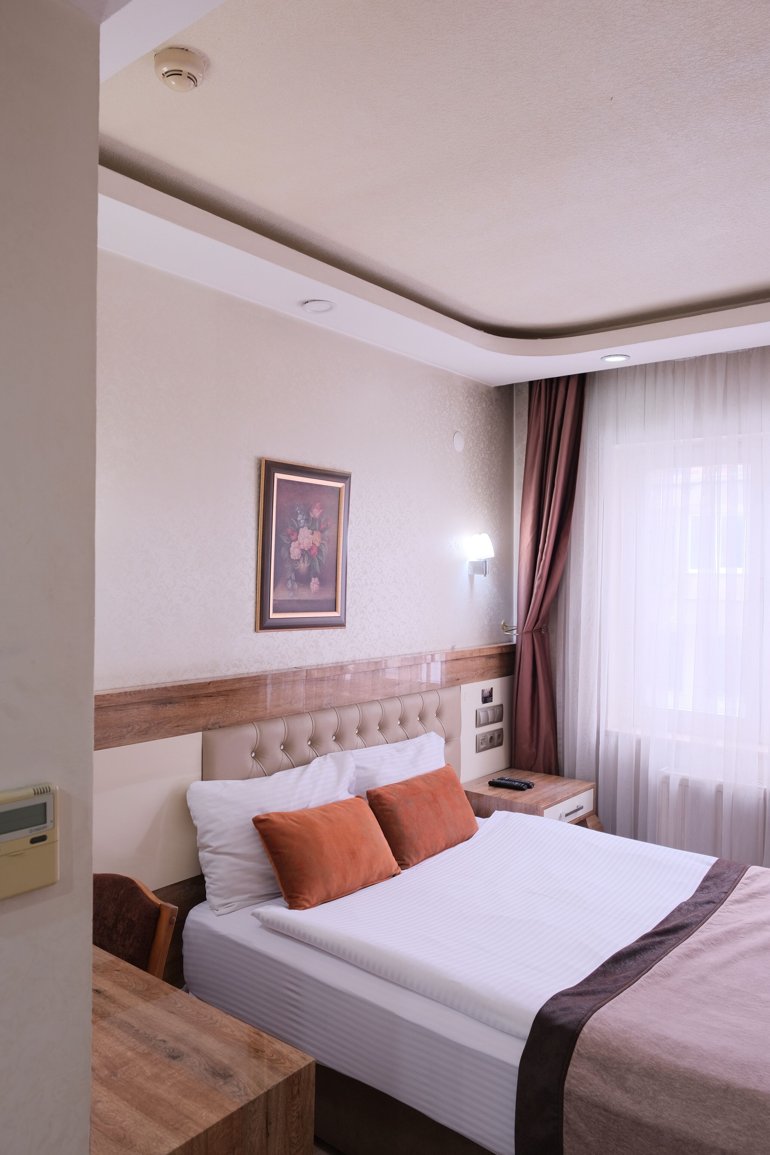 Habitación de hotel con cama, mesita de noche, escritorio de madera, un cuadro en la pared y una ventana con cortinas.