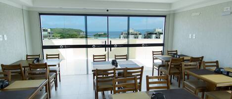Un comedor con mesas y sillas, vista al mar y edificios, y un balcón con puertas de vidrio.