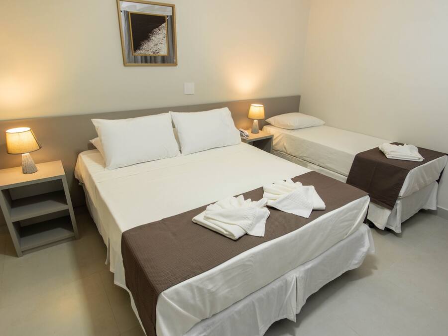 A hotel room with a large bed, two smaller beds, a nightstand with a lamp, and a mirror on the wall.
