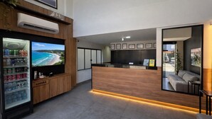 Ein Empfangsbereich mit Holztresen, einem Fernseher, der eine Strandansicht zeigt, einem Getränkeautomaten und einer Couch in der Ecke.
