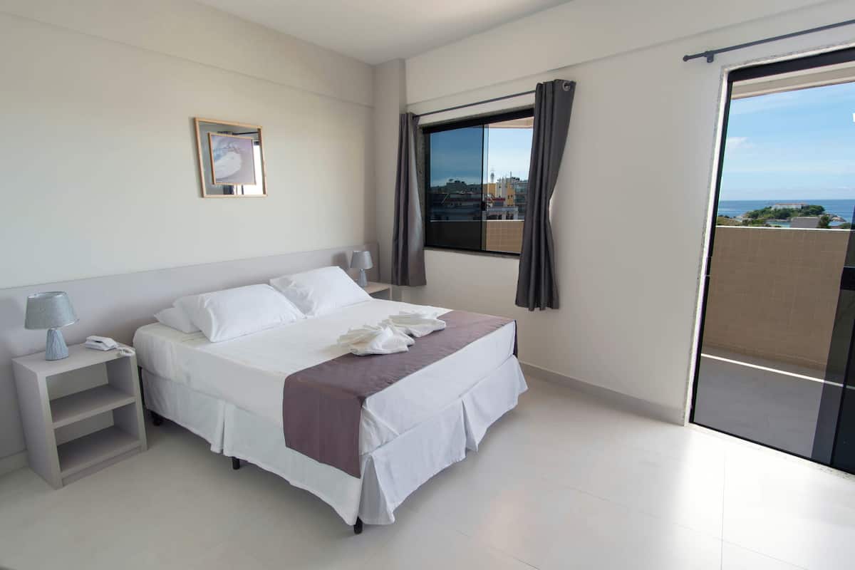 Ein Hotelzimmer mit Bett, Nachttisch, Lampe, einem gerahmten Bild an der Wand und Blick auf die Stadt durch das Fenster.