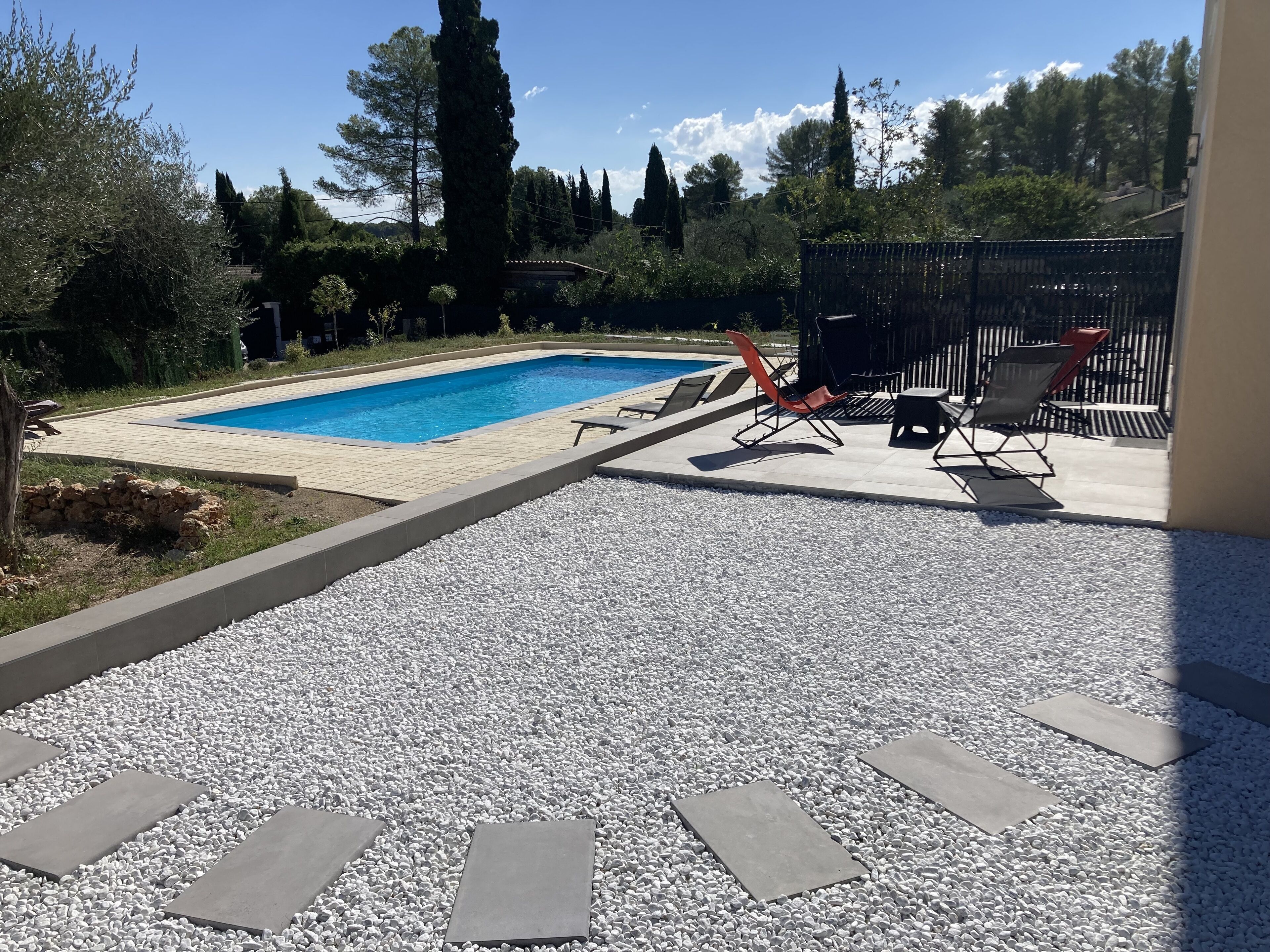 Une piscine rectangulaire avec une terrasse carrelée, entourée de petits galets blancs. On y trouve deux chaises longues et une petite table.